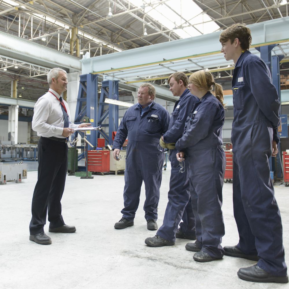 Co-workers in a factory gathered around listening to manager speaking.