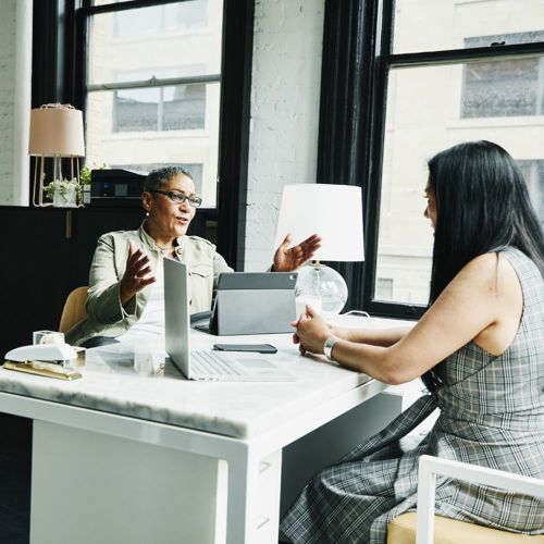 Two people sitting at an office desk with laptop computers open, talking
