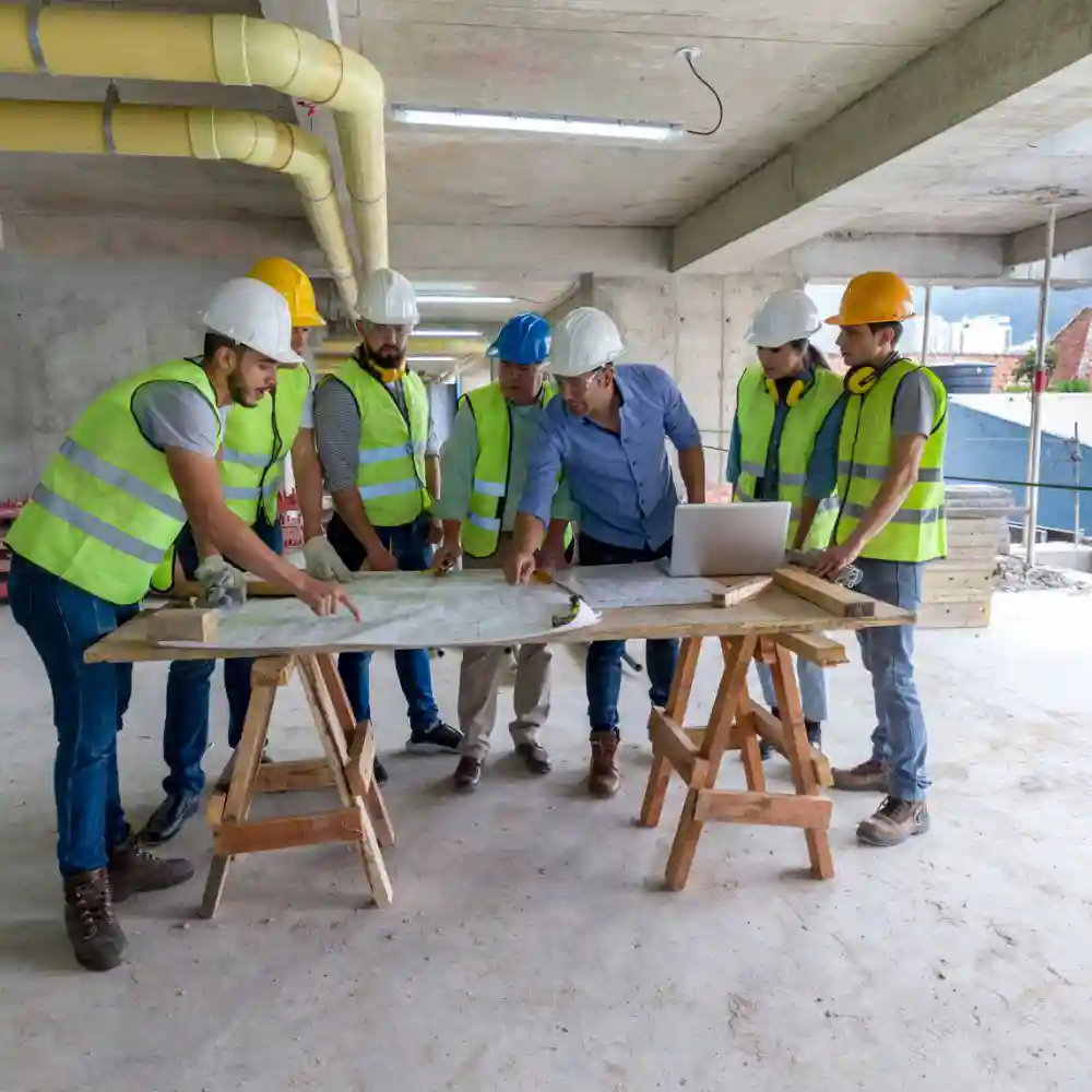 Group of construction workers wearing hard hats, gathered around a table, discussing building plans.