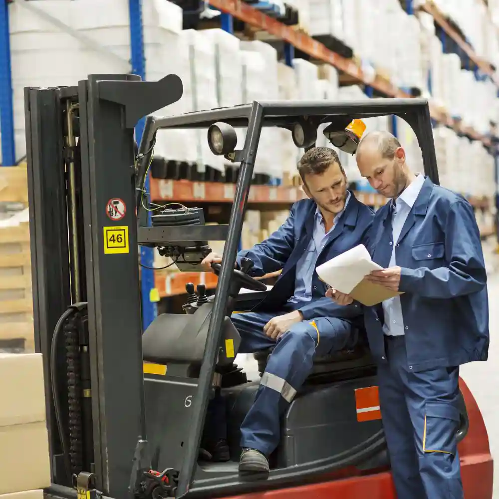 Two people in a warehouse, looking at a document, one person sitting in a forklift.