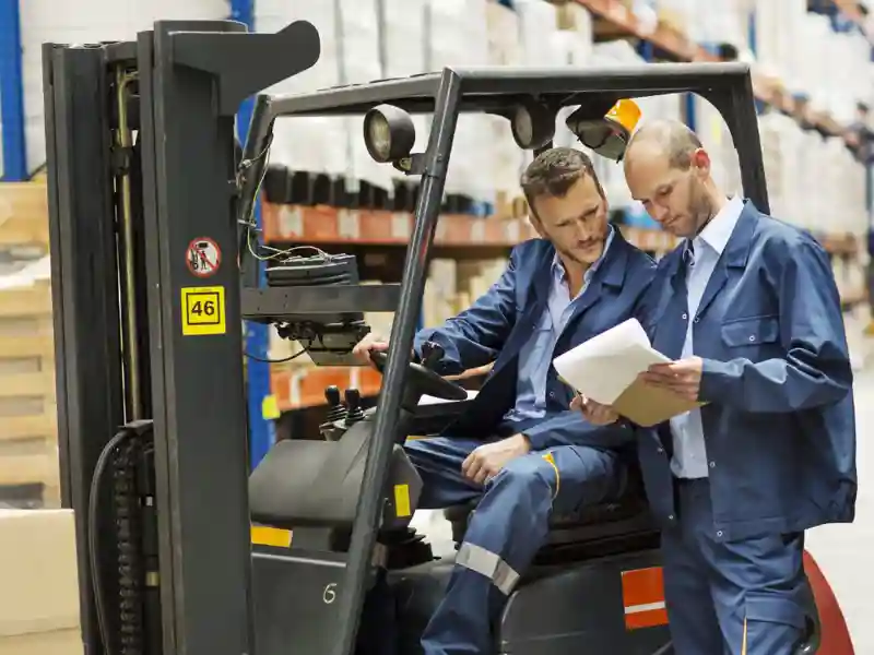 Two people in a warehouse, looking at a document, one person sitting in a forklift.
